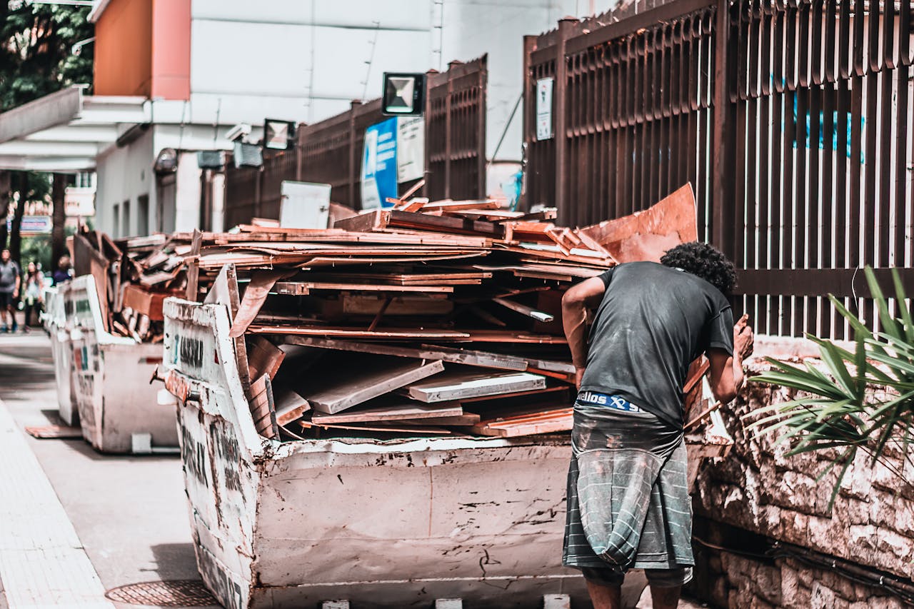 Man collecting materials from a waste container in an urban street, emphasizing recycling and waste management.
